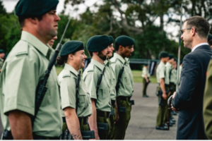 A group of uniformed personnel stand in formation as a suited man speaks with them outdoors.