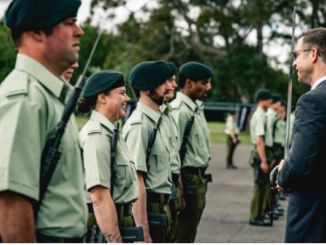 A group of uniformed personnel stand in formation as a suited man speaks with them outdoors.