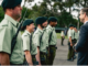 A group of uniformed personnel stand in formation as a suited man speaks with them outdoors.