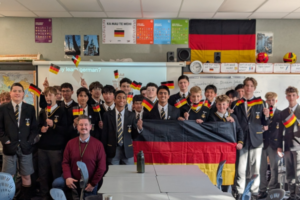 A group of Wellington College students stand together in a classroom holding German flags and a large German banner during a cultural presentation. The students, mostly wearing school blazers and ties, are joined by a teacher seated at the front. German-themed posters and decorations hang on the classroom walls, including a large German flag.
