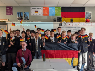 A group of Wellington College students stand together in a classroom holding German flags and a large German banner during a cultural presentation. The students, mostly wearing school blazers and ties, are joined by a teacher seated at the front. German-themed posters and decorations hang on the classroom walls, including a large German flag.