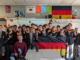 A group of Wellington College students stand together in a classroom holding German flags and a large German banner during a cultural presentation. The students, mostly wearing school blazers and ties, are joined by a teacher seated at the front. German-themed posters and decorations hang on the classroom walls, including a large German flag.