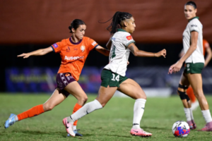 Wellington Phoenix player dribbles forward with the ball while a Brisbane Roar opponent chases closely behind during a semi-final match.