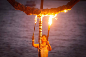 Performer holds two flaming torches beneath a burning ring structure at dusk beside the water.