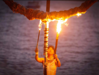 Performer holds two flaming torches beneath a burning ring structure at dusk beside the water.