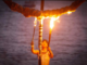 Performer holds two flaming torches beneath a burning ring structure at dusk beside the water.