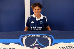Aquila Peleseuma sits in a Paris FC shirt behind a table displaying the club logo and shirts bearing his name.