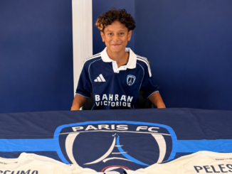 Aquila Peleseuma sits in a Paris FC shirt behind a table displaying the club logo and shirts bearing his name.