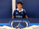 Aquila Peleseuma sits in a Paris FC shirt behind a table displaying the club logo and shirts bearing his name.
