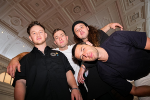 Members of Drax Project pose inside Wellington Town Hall, standing beneath the venue’s ornate restored ceiling ahead of the grand opening concert in February 2027.