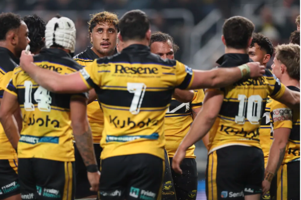 Hurricanes rugby players gather in a tight huddle during a match, wearing yellow and black jerseys. Several players have their arms around teammates as they listen during a stoppage in play, with a packed stadium crowd blurred in the background.