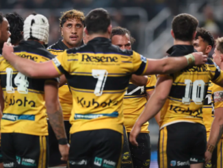 Hurricanes rugby players gather in a tight huddle during a match, wearing yellow and black jerseys. Several players have their arms around teammates as they listen during a stoppage in play, with a packed stadium crowd blurred in the background.
