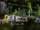 A passenger train sits off the tracks near Khandallah Station in Wellington at night after a derailment. The front carriage is illuminated by artificial light, with trees and overhead power lines visible in the background.