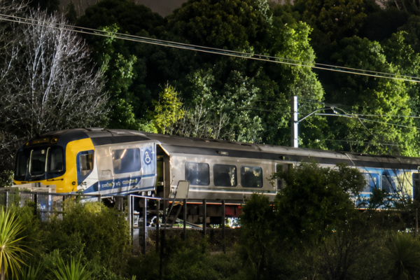 A passenger train sits off the tracks near Khandallah Station in Wellington at night after a derailment. The front carriage is illuminated by artificial light, with trees and overhead power lines visible in the background.