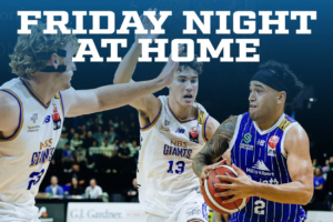 A Wellington Saints player drives with the ball against two Nelson Giants defenders, with “Friday Night at Home” text across the image.