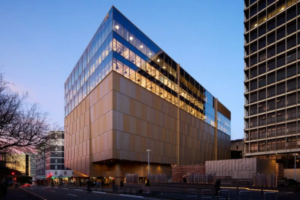 Te Rua Archives New Zealand building in Pipitea, Wellington, viewed at dusk. The large modern archive facility features a copper-toned lower façade and glass upper levels glowing with interior lights, with pedestrians and traffic visible along the street.