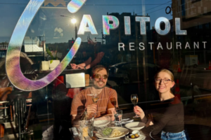 Two diners raise glasses inside Capitol Restaurant in Wellington, seated by the front window beneath the restaurant's logo with dishes from the Wellington On a Plate menu on the table.