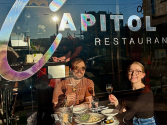 Two diners raise glasses inside Capitol Restaurant in Wellington, seated by the front window beneath the restaurant's logo with dishes from the Wellington On a Plate menu on the table.