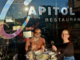 Two diners raise glasses inside Capitol Restaurant in Wellington, seated by the front window beneath the restaurant's logo with dishes from the Wellington On a Plate menu on the table.