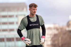 A goalkeeper in a green training shirt and gloves stands on a football field with blurred city buildings behind him.
