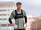 A goalkeeper in a green training shirt and gloves stands on a football field with blurred city buildings behind him.