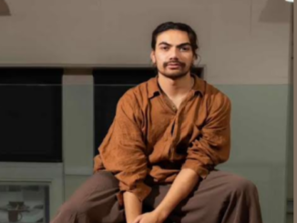 Kasi Valu seated indoors, wearing a rust-brown shirt and dark trousers.