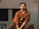 Kasi Valu seated indoors, wearing a rust-brown shirt and dark trousers.