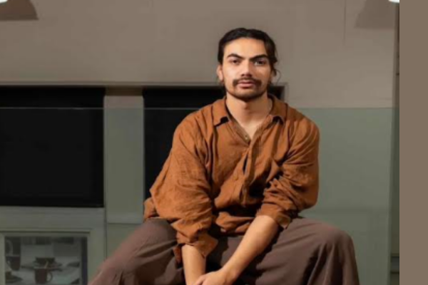 Kasi Valu seated indoors, wearing a rust-brown shirt and dark trousers.