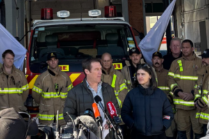 Two people speak to media in front of a fire engine, with a group of firefighters standing behind them inside a station.