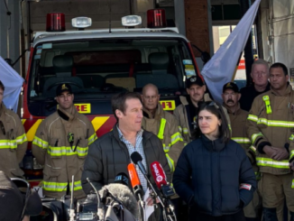 Two people speak to media in front of a fire engine, with a group of firefighters standing behind them inside a station.