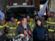 Two people speak to media in front of a fire engine, with a group of firefighters standing behind them inside a station.