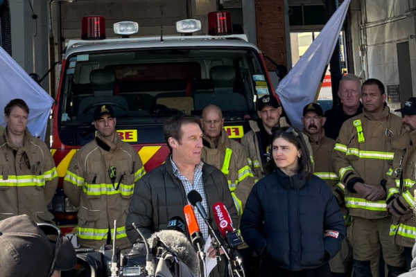 Two people speak to media in front of a fire engine, with a group of firefighters standing behind them inside a station.