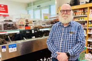 A bearded man in glasses stands inside a deli beside a glass counter filled with food and shelves of grocery products.