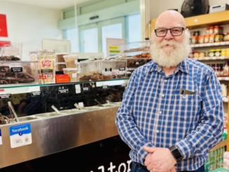 A bearded man in glasses stands inside a deli beside a glass counter filled with food and shelves of grocery products.