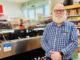 A bearded man in glasses stands inside a deli beside a glass counter filled with food and shelves of grocery products.