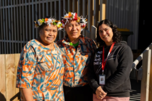 Three people stand together outside a wooden building, with two wearing matching patterned outfits and flower head garlands.