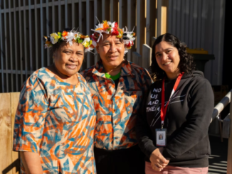 Three people stand together outside a wooden building, with two wearing matching patterned outfits and flower head garlands.