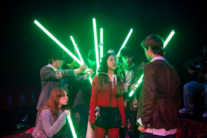 Wellington College pupils perform a dramatic scene from Make Your Mirror Be Me, surrounded by glowing green light tubes.
