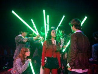 Wellington College pupils perform a dramatic scene from Make Your Mirror Be Me, surrounded by glowing green light tubes.