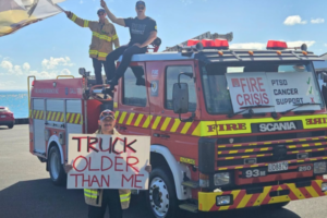 Firefighters stand on and beside an older fire engine holding signs about ageing appliances, health risks and the fire service crisis.