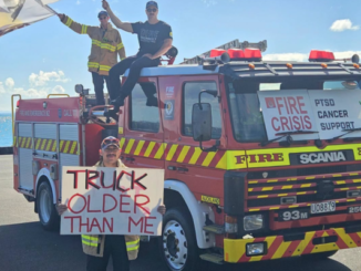 Firefighters stand on and beside an older fire engine holding signs about ageing appliances, health risks and the fire service crisis.