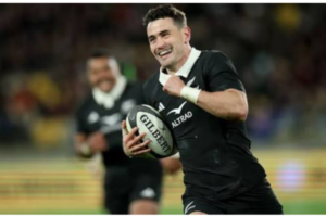 An All Blacks player smiles while running with the ball during a rugby match, with a teammate blurred in the background.