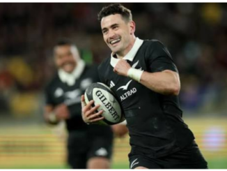 An All Blacks player smiles while running with the ball during a rugby match, with a teammate blurred in the background.