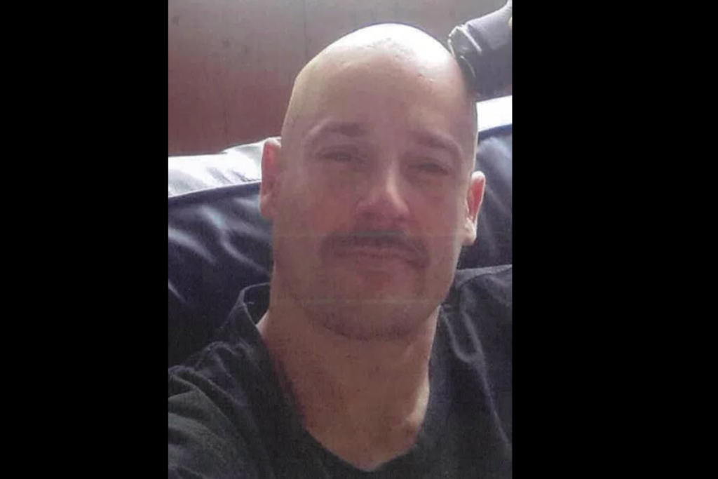 A close-up photo of a man with a shaved head and moustache, wearing a black top and seated indoors.