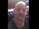 A close-up photo of a man with a shaved head and moustache, wearing a black top and seated indoors.