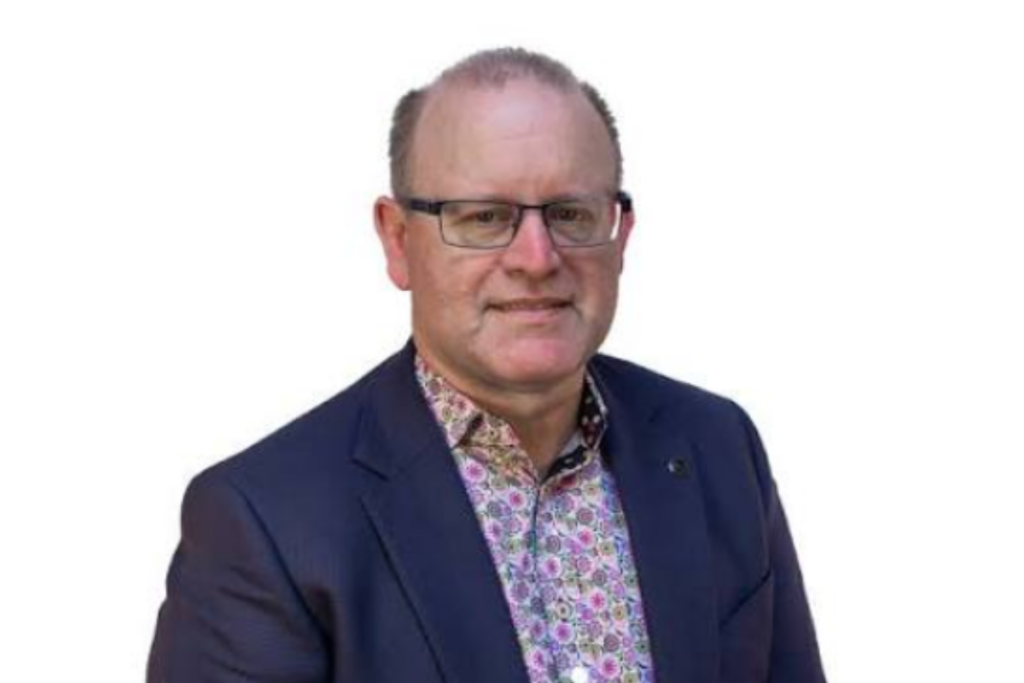 A man wearing glasses, a navy blazer and a patterned shirt poses against a white background.