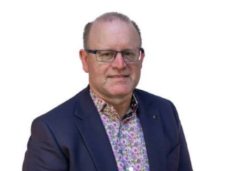 A man wearing glasses, a navy blazer and a patterned shirt poses against a white background.