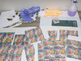 A workbench covered with colourful printed sheets and small cards, alongside gloves, a cutting mat, tools and a spray bottle.