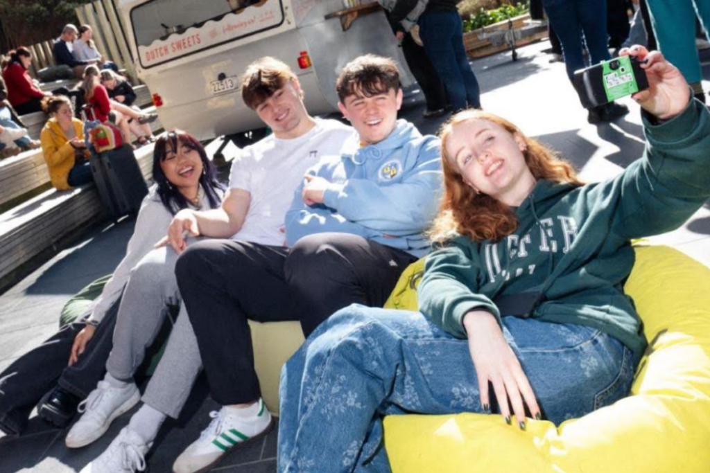 Four young people relax together on colourful beanbags outdoors, with one holding up a camera for a group selfie.