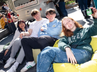 Four young people relax together on colourful beanbags outdoors, with one holding up a camera for a group selfie.